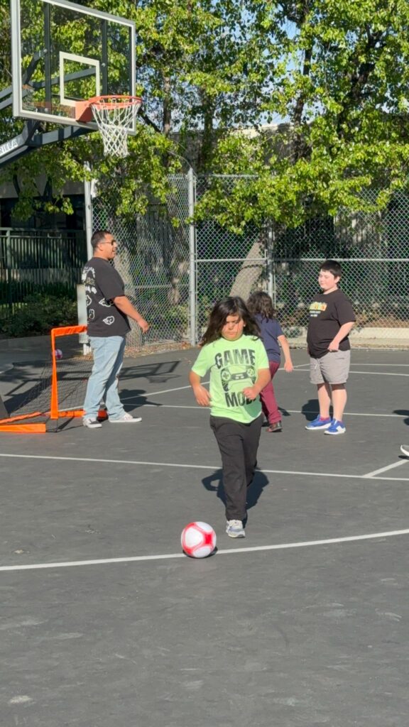 Young kids playing soccer with teacher outside on school playground.